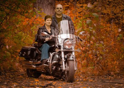 “Couple in their forties pose with a large motorcycle in a wooded setting, captured during a lifestyle portrait session by David Thielen Photographic Artistry”