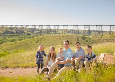 “Family poses together on top of the river valley in Lethbridge with the High Level Bridge spanning the background, capturing the full valley rather than the typical river-bottom view with just steel girders or pylons, photographed by David Thielen Photographic Artistry”