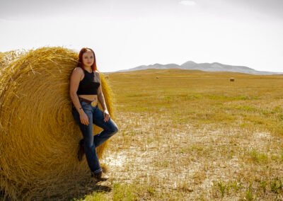 “Graduation portrait of a young woman dressed in jeans and a crop top, leaning against a large round bale with the Sweetgrass Hills in the background at Writing-On-Stone Provincial Park, captured by David Thielen Photographic Artistry”