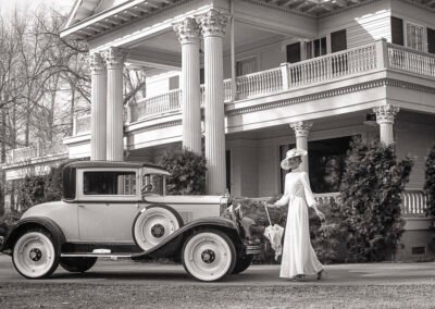 “Model in a long flowing 1930s-style dress with parasol and large hat walks toward a restored 1930s classic car in front of Norland Estates, a historic southern colonial building, captured by David Thielen Photographic Artistry”