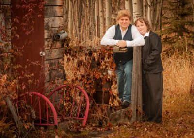 “Couple poses together leaning on a wooden fence attached to an old shed in a landscaped outdoor setting, captured by David Thielen Photographic Artistry”
