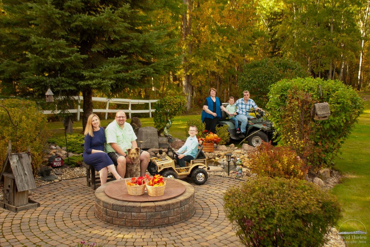 Extended-Family-Backyard-Portrait-in-Alberta An extended family poses for a portrait in their backyard in Alberta. The group includes Grandpa, Grandma, a husband and wife, and their two children, all smiling together in front of a backdrop of trees