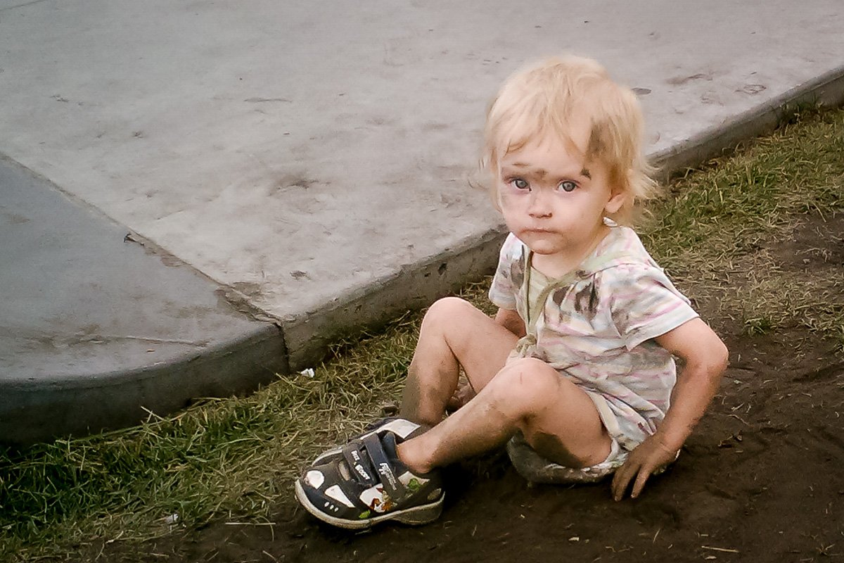 Getting-Dirty-It's-a-talent A young child, freshly bathed, sits in a patch of dirt in an otherwise grassy park, looking up with a mix of curiosity and calm. Her clothes and skin are smudged with dirt, adding charm and realism to the moment.