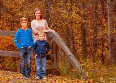 A mother stands with her two sons in front of a wooden fence, surrounded by autumn leaves and fall colors in a warm outdoor family portrait.
