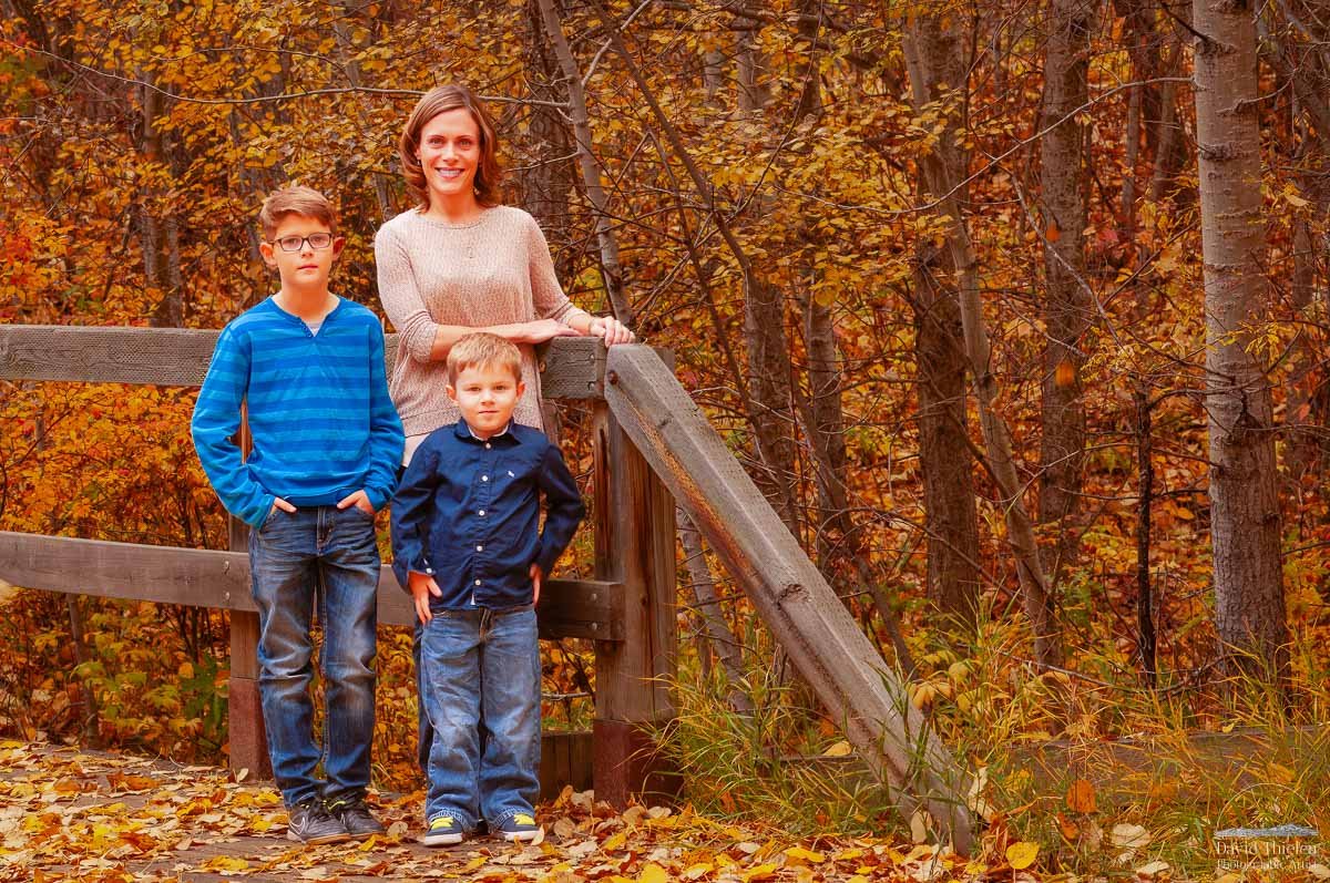 Lethbridge-family-photography A mother stands with her two sons in front of a wooden fence, surrounded by autumn leaves and fall colors in a warm outdoor family portrait.