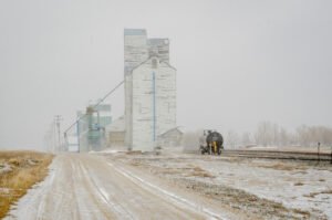 Historic wooden grain elevators in Warner, Alberta, disappear into a late spring snowstorm. A gravel road leads toward the vanishing structures, while an abandoned rail tanker car rests on the tracks, adding to the scene’s eerie atmosphere.